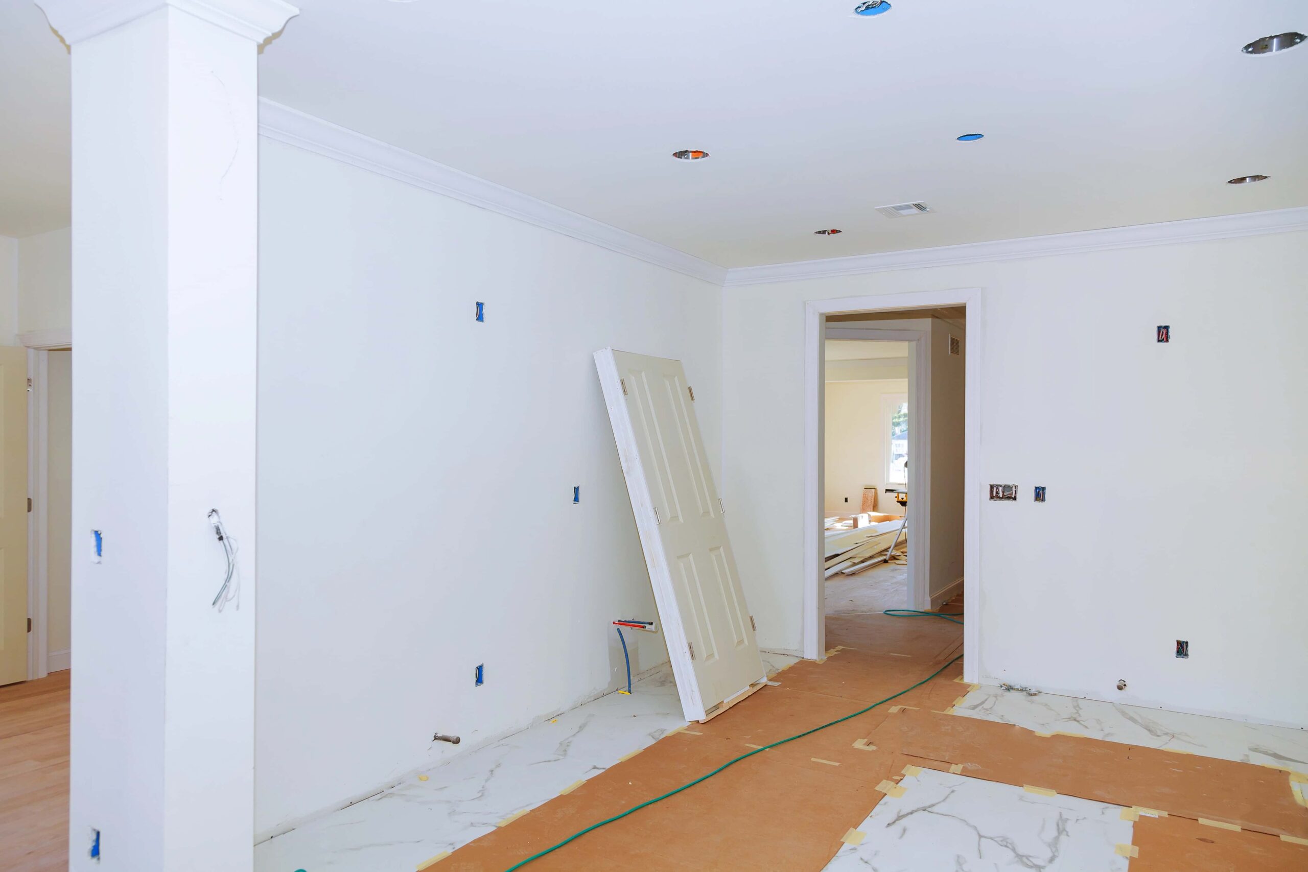 Ein weiß gestrichener Raum im Umbau mit offener Tür, abgedecktem Boden und sichtbaren Elektroanschlüssen an den Wänden.